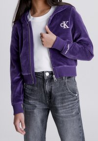 Fialová sametová bunda na zip s kapucí, s bílým logem na hrudi a žebrovanými manžetami, v kombinaci se šedými džíny.