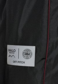 Sorte stofjakke med en grå label med prægede tekst og logo. Pyntet med mørkerødt pipings langs kanten.