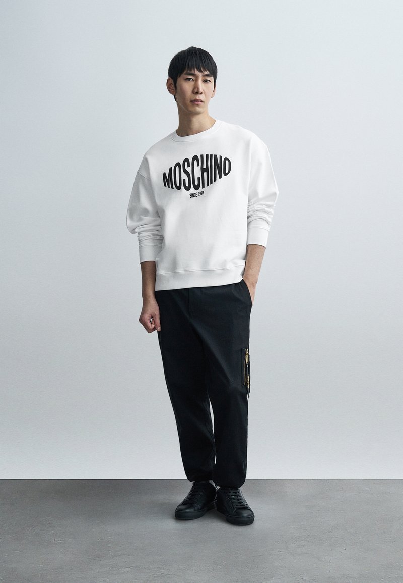 Sweatshirt blanc avec logo "MOSCHINO" noir, associé à un pantalon noir avec une poche zippée. Des baskets noires complètent la tenue.
