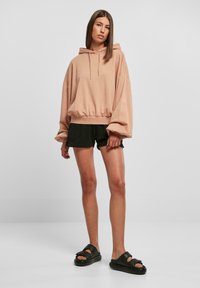 Üks suur roosakas kapuutsiga sviiter, elastse alumise serva, nööriga ja laiade varrukatega, koos mustade lühikeste püksidega ja jämedate mustade sandaalidega heledal taustal.