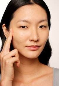 Modella con pelle liscia e chiara e capelli neri lisci. Indossa un top grigio e tocca delicatamente la guancia con una mano, sfoggiando un trucco naturale.