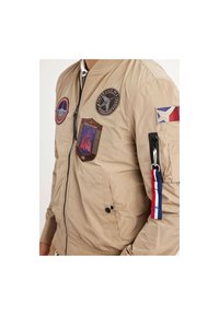 Veste bomber beige avec des écussons, poche zippée sur la manche, poignets et col côtelés. Présente divers motifs brodés et textures.