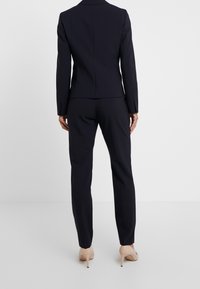 Costume bleu marine avec un blazer ajusté et un pantalon cintré, fabriqué dans un tissu lisse. Le blazer a un dos structuré et le pantalon est fuselé.