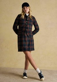 Marynarka i spódnica w kratę w kolorach granatowym, zielonym i czerwonym, z złotymi guzikami i kieszeniami; w połączeniu z białymi skarpetkami i granatowymi trampkami.