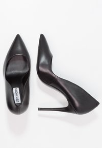 Pár černých kožených lodiček na vysokém podpatku se špičatou špičkou, jedna bota je zachycena z vrchu, ukazuje vnitřní označení „Steve Madden“, druhá z bočního pohledu.