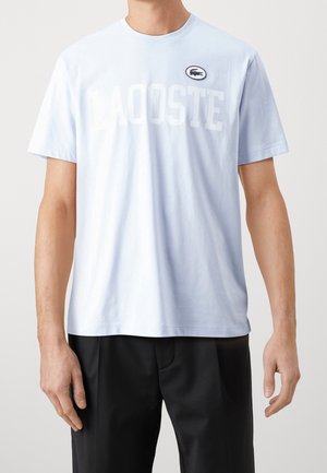 Mies, jolla on vaaleansininen lyhythihainen Lacoste-paita ja mustat housut, seisoo yksinkertaista vaaleaa taustaa vasten.