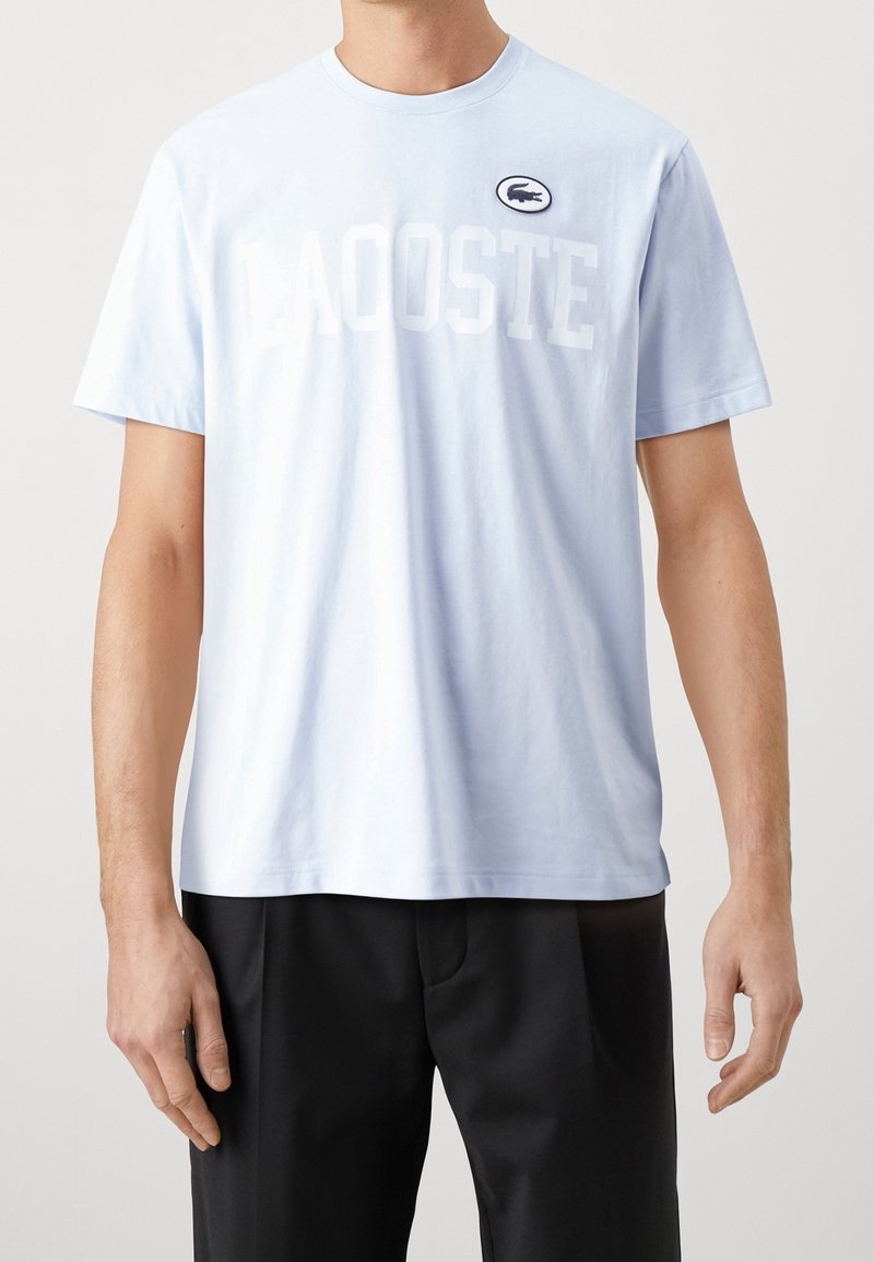 Homme portant un t-shirt Lacoste bleu clair à manches courtes et un pantalon noir, debout devant un fond clair uni.