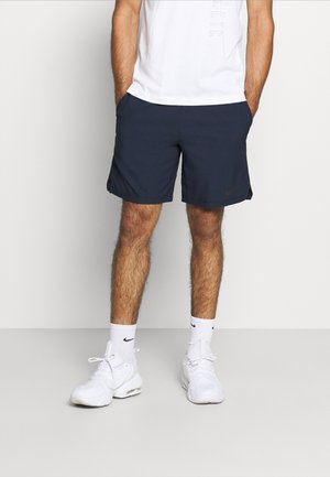 Man die marineblauwe sportshorts draagt, witte sportsokken met een zwart logo, witte sneakers en een wit T-shirt, staand met handen in de zakken.