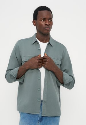 Uomo nero che sistema una camicia verde chiaro sbottonata sopra una maglietta bianca, indossa jeans blu, in piedi davanti a uno sfondo chiaro e uniforme.