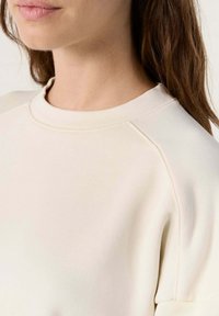 Felpa beige chiaro con collo rotondo classico, maniche raglan e una superficie liscia. Dettagli di cucitura lungo il collo e le cuciture delle spalle.