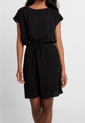 Femme portant une courte robe noire cintrée à la taille avec des manches courtes sur un fond clair uni.