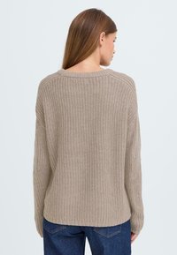 Beiger Strickpullover mit weitem Schnitt, weitem Ausschnitt und gerippter Textur. Lange Ärmel mit überschnittenen Schultern, kombiniert mit dunkelblauen Jeans.