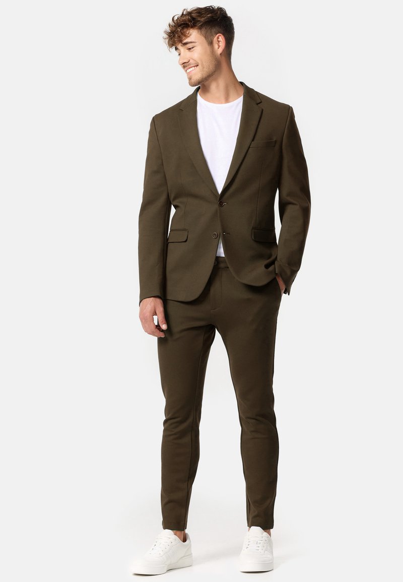 Completo verde oliva con giacca slim-fit e pantaloni, dotato di chiusura con un solo bottone e tasche, abbinato a una maglietta bianca e sneakers bianche.