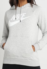 Harmaa huppari, joka on valmistettu pehmeästä kankaasta. Siinä on suuri valkoinen Nike-logo edessä ja kengurupocket.