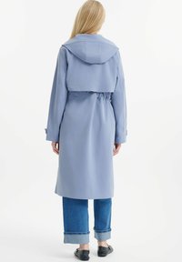 Hellblauer Trenchcoat mit Kapuze, tailliert und langen Ärmeln. Der Rücken hat eine kurze Schicht und wird mit blauen Jeans und schwarzen Schuhen kombiniert.