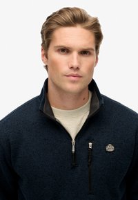 Marineblaues Zip-Up-Pullover aus strukturiertem Stoff, mit hohem Kragen und kontrastierenden dunkelgrauen Akzenten. Mit einem Logo-Patch auf der linken Brust.