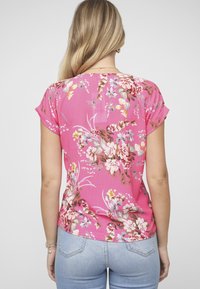 Top floral de manga corta en rosa brillante, con diversos patrones de flores en blanco, amarillo y verde, confeccionado en una tela ligera y texturizada.