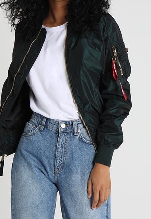 Personne portant une veste bomber noire avec un t-shirt blanc et un jean en denim bleu taille haute sur fond uni.