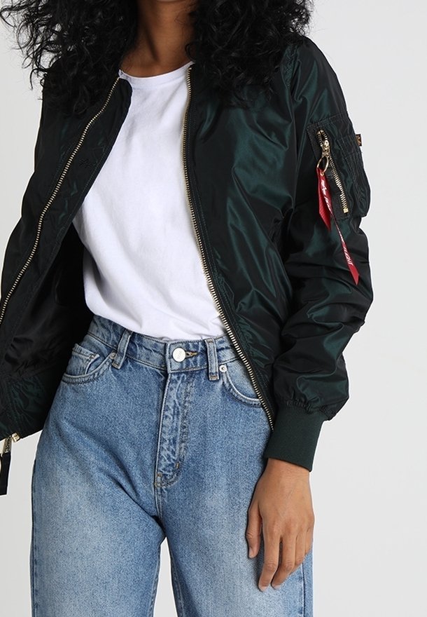 Personne portant une veste bomber noire avec un t-shirt blanc et un jean en denim bleu taille haute sur fond uni.
