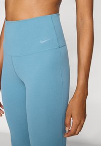 Visoke plave tajice izrađene od rastezljivog materijala, s malim sivim Nike logotipom na kuku i ravnim šavovima za udobnost.