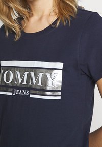 Námořnická modrá bavlněná tričková s kulatým výstřihem, s kovově stříbrným logem "TOMMY JEANS" uvnitř bílého obdélníkového rámečku.