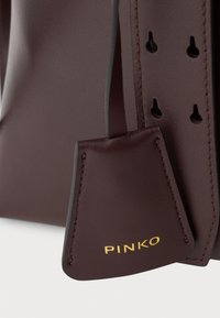 Tmavě burgundská kožená taška s hladkým povrchem, se zlatým logem "PINKO" a zesíleným prošíváním. Obsahuje otvory pro nastavitelné popruhy.