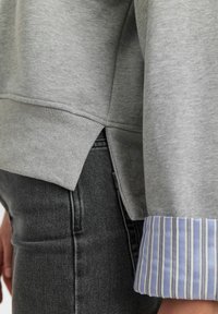 Grå sweatshirt med ribbad nederkant och sidoflikar, matchad med mörka jeans. Ärmarna har blåa och vita ränder som ger kontrast.