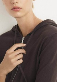 Felpa con zip di colore viola scuro presenta una texture morbida con cordini e una cerniera argentata. La mano del modello si appoggia sul tessuto vicino alla cerniera.