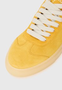 Sneaker giallo in suede con una texture morbida, caratterizzato da lacci bianchi e una suola in gomma traslucida. Dettagli di cucitura curva lungo la tomaia.