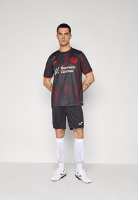 Mørkegrå fodboldtrøje med røde accenter og logo, parret med sorte shorts. Hvide skinnebeskyttere og støvler fuldender outfittet.