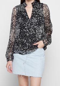 Schwarze Blumenshirt mit transparenten langen Ärmeln, V-Ausschnitt, gerafften Details an den Schultern, getragen mit einem hellen Jeansrock.