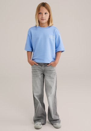 Hellblaues, oversized T-Shirt mit weißem grafischem Text auf der linken Brust, kombiniert mit grauen Schlagjeans und grauen Sneakers.
