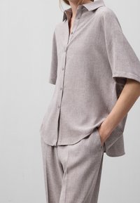 Ljust grå kortärmad skjorta med knäppning framtill, tillverkad av textil med struktur. Inkluderar en klassisk krage och sidoklyftor för ökad rörelsefrihet.