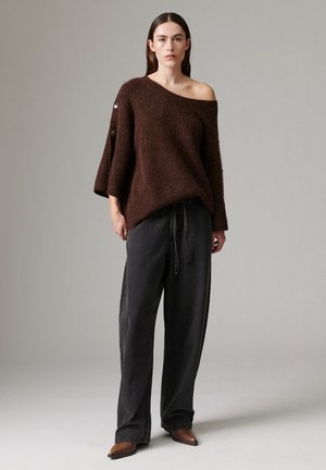 Femme portant un pull marron ample décolleté, un pantalon large noir avec cordon de serrage et des chaussures marron pointues, debout devant un fond gris.