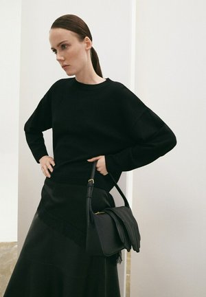 Mujer con cabello oscuro recogido, vestida con suéter y falda negras, sosteniendo un bolso negro con guantes colgados, de pie frente a paredes claras.