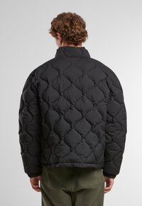Chaqueta de plumas negra acolchada con cuello alto. Presenta un patrón de diamantes texturizado y puños elásticos para un ajuste ceñido. Costuras visibles como acentos.
