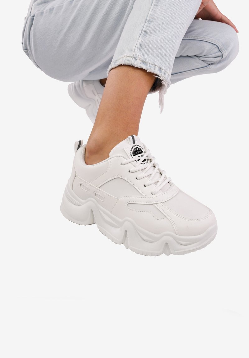 Zapatillas blancas gruesas de cuero sintético, con punteras redondeadas, suelas gruesas y cortes de ventilación a los lados. Detalle del logo negro en la lengüeta.