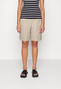 Personne portant un short beige jusqu'aux genoux, un haut à rayures noir et blanc, et des chaussures à enfiler noires à bout ouvert, se tenant sur un fond uni.