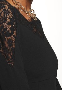 Svart klänning med spetsdetaljer på axlarna och i halsringningen, med en slät tygkänsla och ett kraftigt guldhalsband.