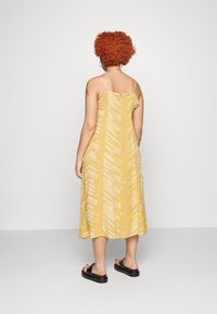 Vestido maxi amarelo com padrão de zigue-zague, com alças finas e um corte descontraído. Usado com sandálias slide pretas.