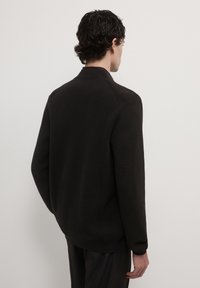 Homme avec des cheveux bouclés foncés portant un pull à manches longues noir et un pantalon noir, tourné le dos contre un fond clair uni.