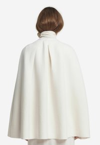 Biała, teksturowana wełniana peleryna z wysokim kołnierzem, z centralnym zakładką z tyłu dla dodania objętości. Minimalistyczny design, bez widocznych elementów metalowych ani akcentów.