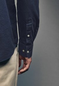 Námornícka košeľa s gombíkmi, vyrobená z texturovanej látky, s špičatým manžetovým koncom so dvoma gombíkmi a trojuholníkovým detailom podpažia pri lakti.