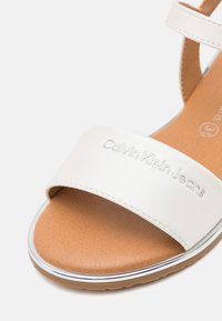 Sándalo blanco de Calvin Klein Jeans con plantilla marrón, ribete plateado en el borde de la suela y correa ajustable en el tobillo.