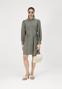 Rochie scurtă khaki cu mâneci bufante, cu deschidere cu butoni în față, talie plisată și textură netedă; asortată cu o geantă de umăr crem și sandale.