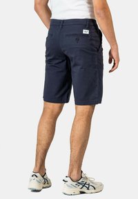 Navy blue cotton shorts with a straight cut, featuring rear pockets, button details, and visible stitching. Worn with white and navy sneakers.