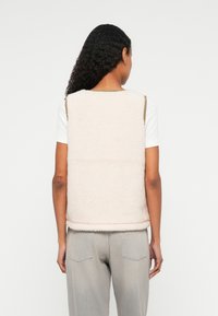 Gilet en polaire beige clair avec une texture douce, un col rond et des finitions brunes contrastantes. Stylé avec une chemise blanche et un pantalon gris.