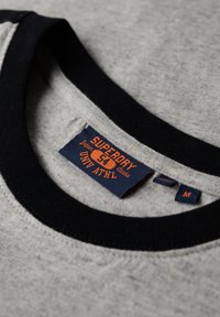 Grå t-shirt med en sort ribbet krave. Har et blåt mærke med orange tekst, der står "SUPERDRY," som angiver størrelse og mærkeoplysninger.