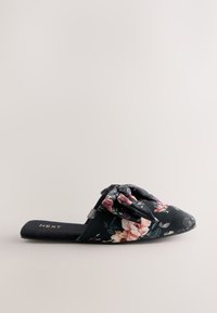 Zapatilla sin cordones con estampado floral sobre un fondo oscuro, con un lazo anudado como adorno y suela plana y acolchada. Logo de la marca en la plantilla.