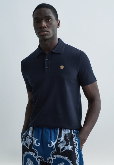 Navy polo shirt features a three-button placket and a gold embroidered logo. Paired with patterned blue and black shorts.